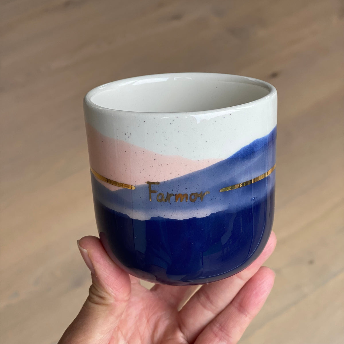 Farmor kop i håndlavet keramik med kongeblå og blush glasur, guldbånd og hvid inderside, holdt i hånden.