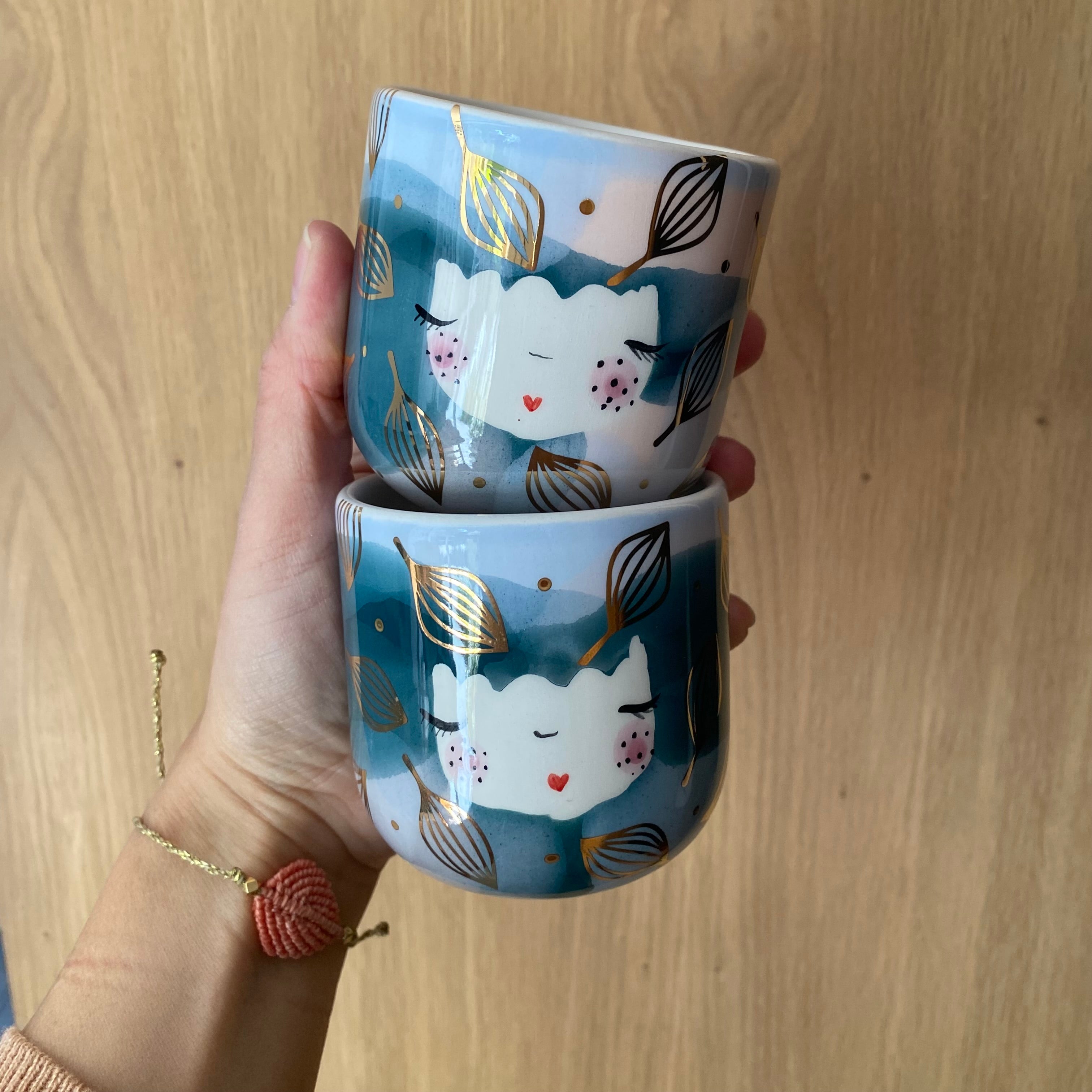 To cappuccinokopper med guldblade og blå og lyserøde farver, holdt i hånden – håndlavet keramik med naturinspirerede detaljer