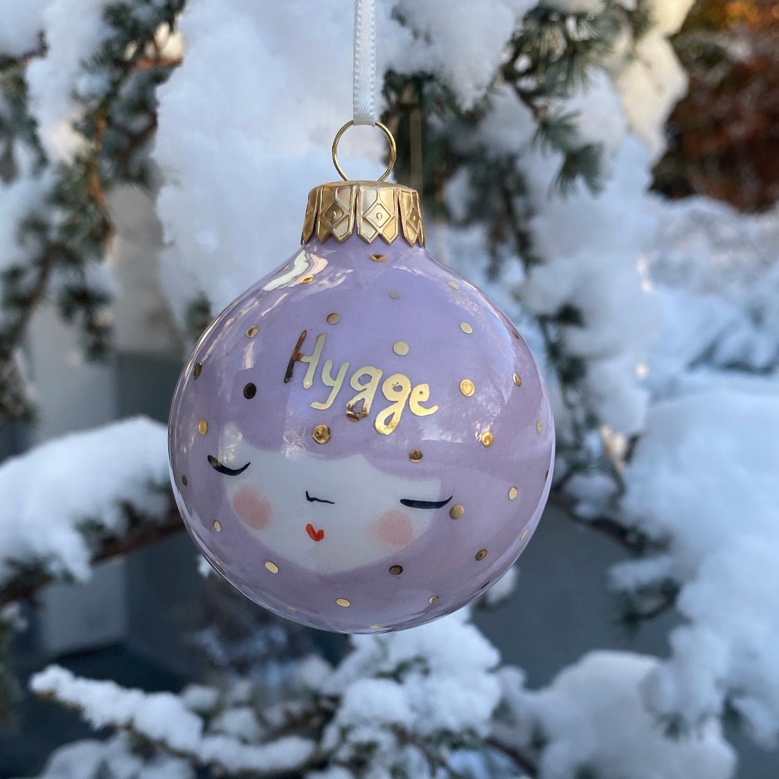Lyselilla julekugle med teksten Hygge i guld, hænger på et juletræ med sne – håndlavet keramik fra unika:k.