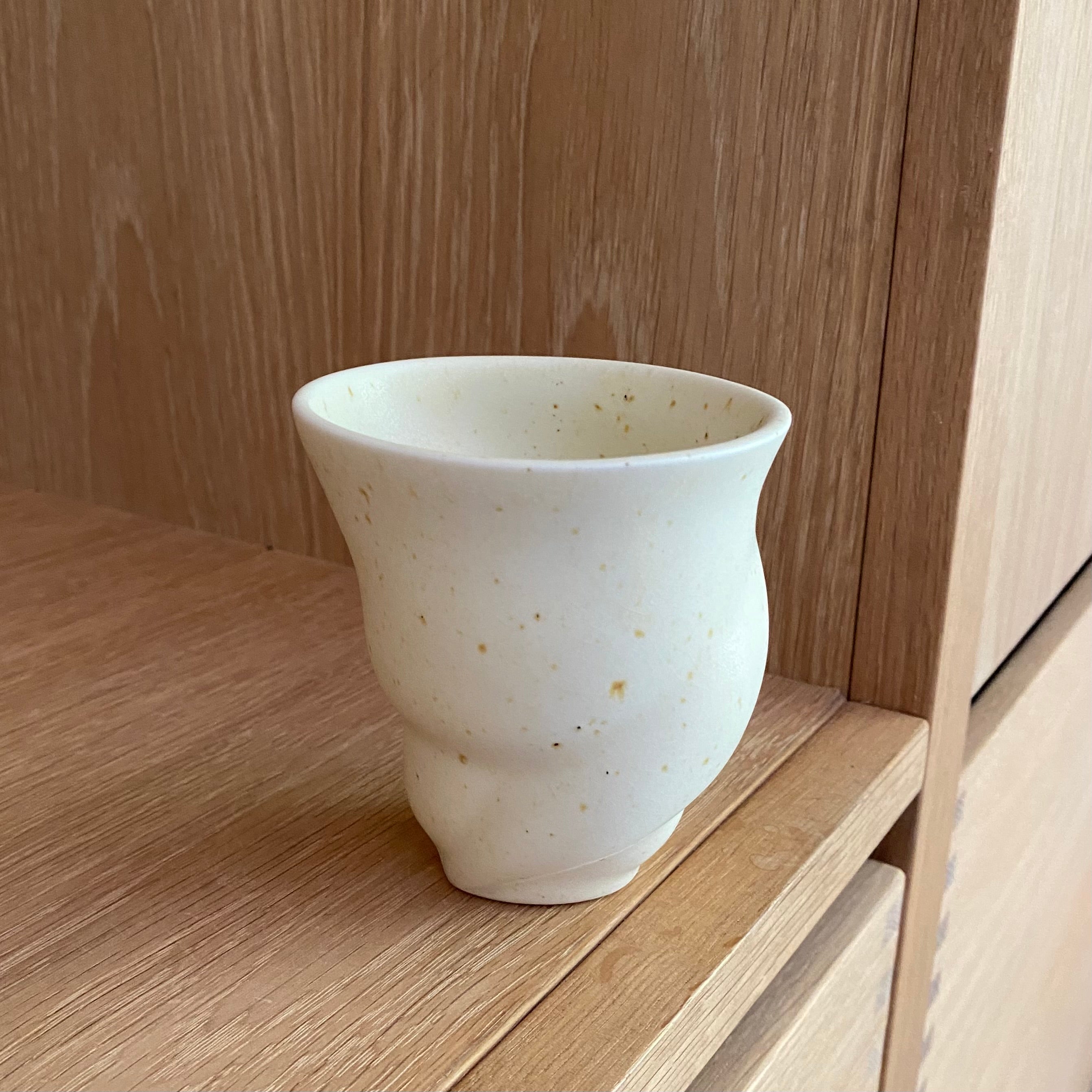 “Skal”-kaffekop i lys, håndlavet porcelæn med pletter – placeret på keramikhylde