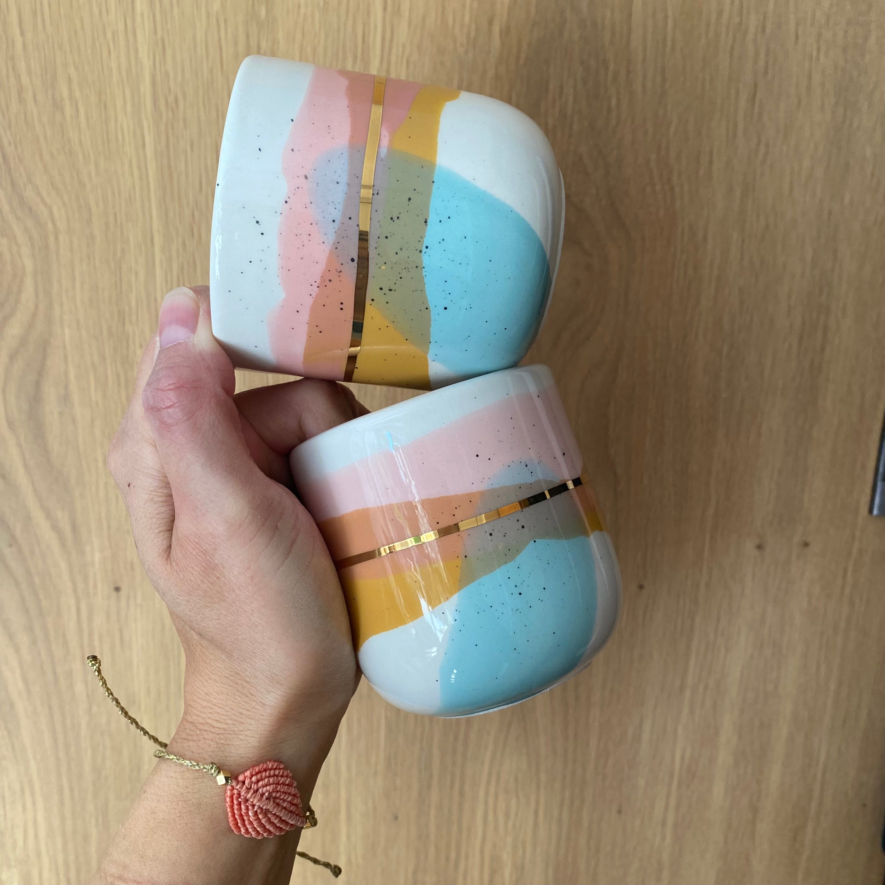 To kaffekopper “Landscape” holdt i hånden – håndlavet keramik med lyserød, turkis, gul og guld fra Marinski Heartmades