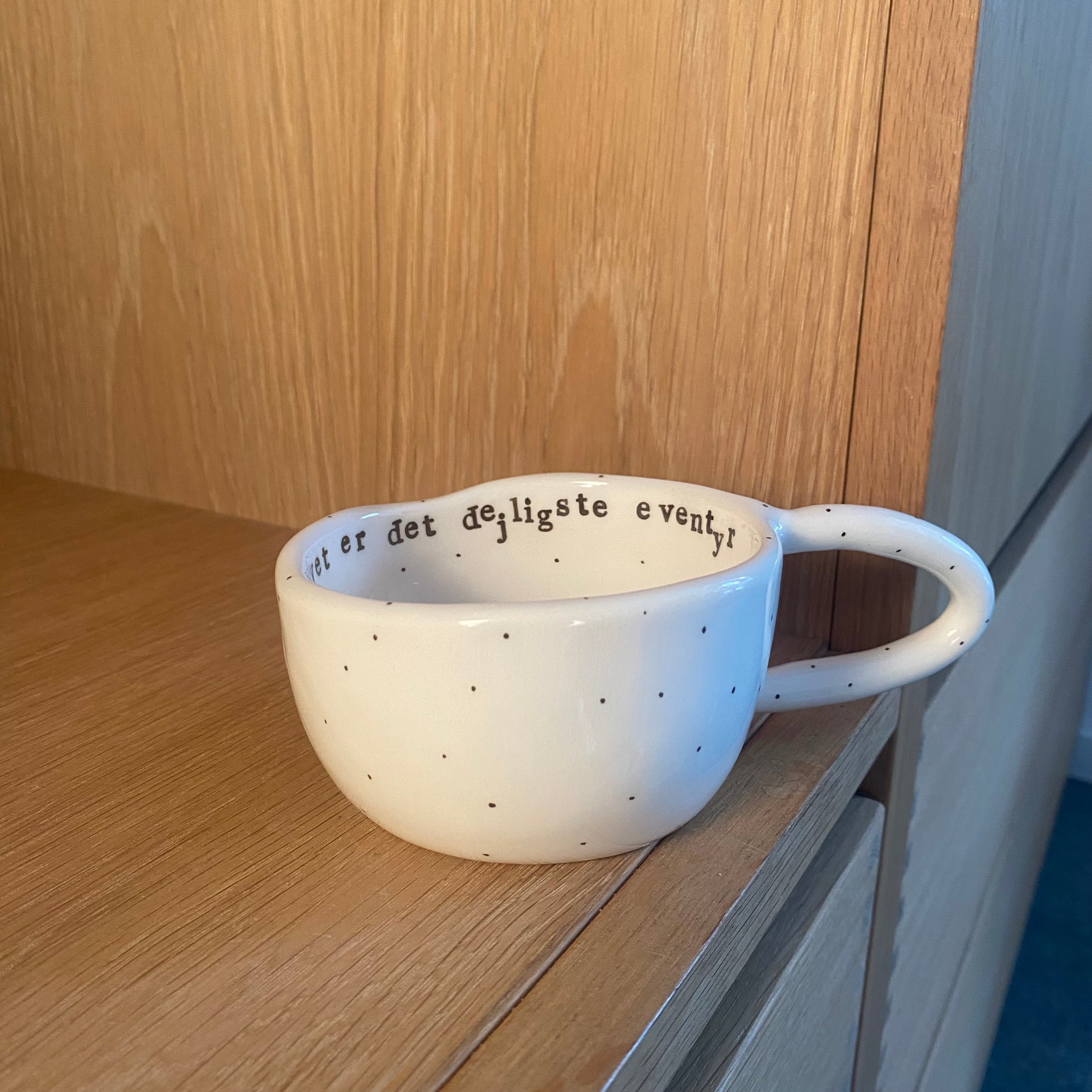 Håndlavet kop med hank og teksten “Livet er det dejligste eventyr” i hvid keramik med sorte prikker