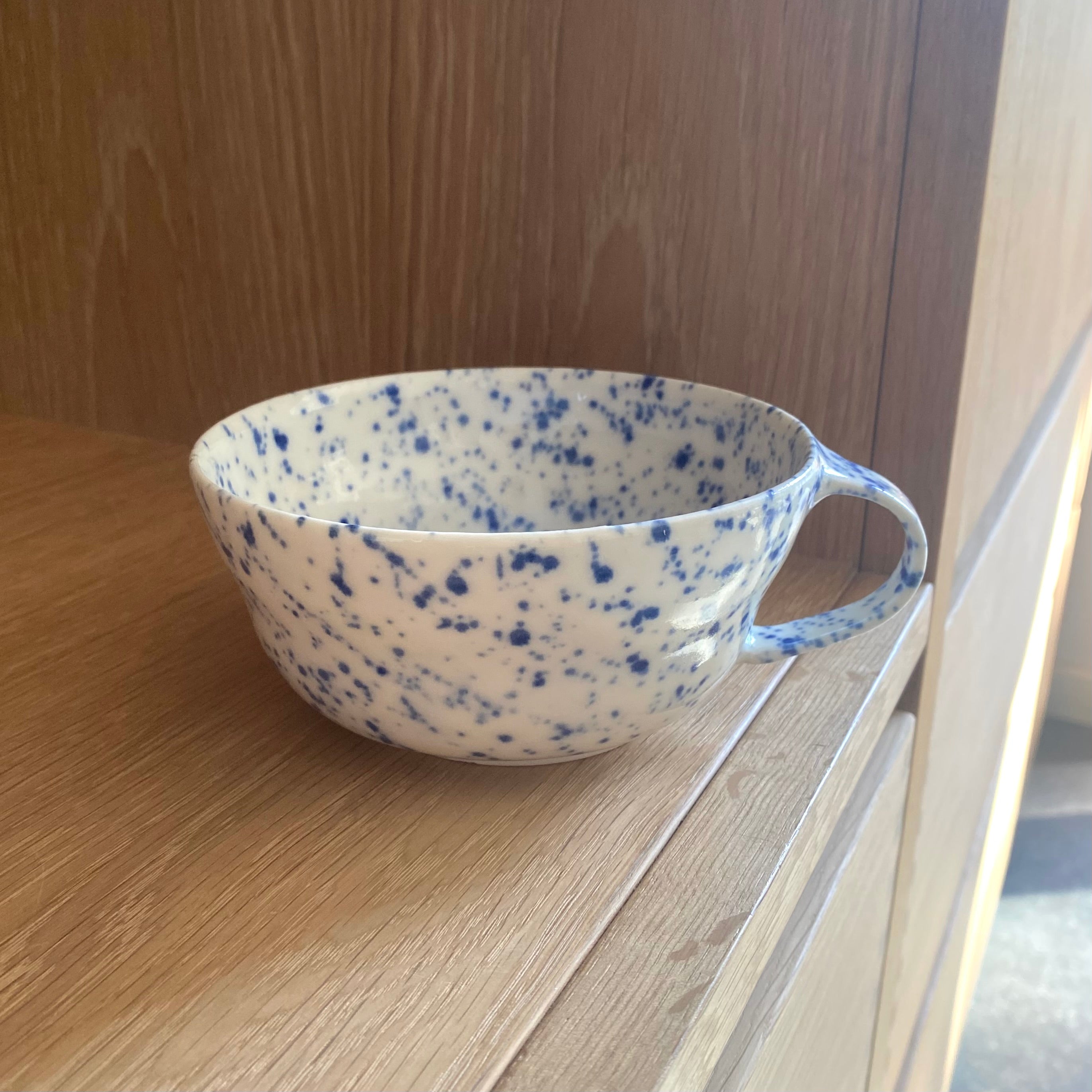 Håndlavet tekop i porcelæn med blå splash, står på bord.